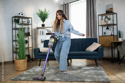 Attractive woman in headphones listening music and doing housekeeping using modern wireless vacuum cleaner. Young housewife enjoying cleaning time with modern technology. photo