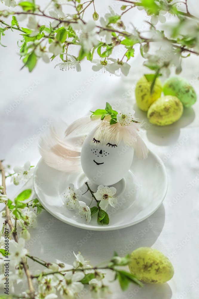 Happy easter. Natural egg with painted cute face in floral wreaths on linen fabric with blooming spring branch, petals and green leaves in soft white light. Creative Easter background.