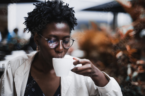 Portrait of a young glamorous black female in classy spectacles drinking a hot tea in a street cafe; ravishing African woman in glasses drinking a delicious latte from the cup in an outdoor restaurant