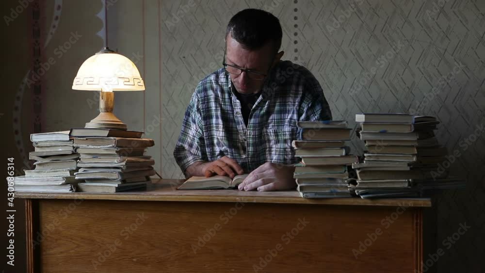 A man sits at a table with many books. A man with glasses reads a book and looks thoughtfully out the window.