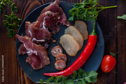 Pieces of meat, pork and chicken, with herbs and paprika and tomatoes. Traditional Ukrainian snacks, cutting board, cold smoked meat cutting on a black plate. View from above. 