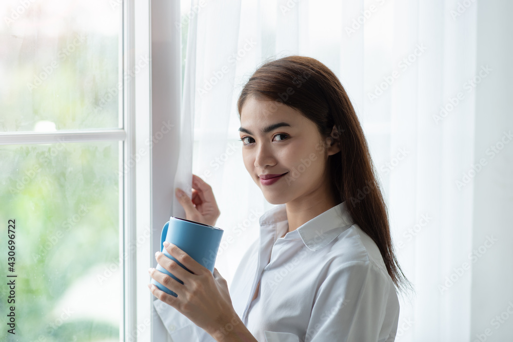 Asian woman drinking a cup of coffee and looking through the glass window after wake up in the morning Asian woman drinking a cup of coffee and looking through the glass window after wake up in the morning