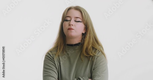 Pretty blonde long haired woman in green sweatshirt with crossed arms looks disgustedly at camera by white wall slow motion photo