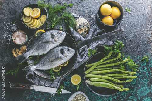 Fresh uncooked dorado or sea bream fish with lemon ,asparagus, herbs, olive oil and spices over black backdrop, top view