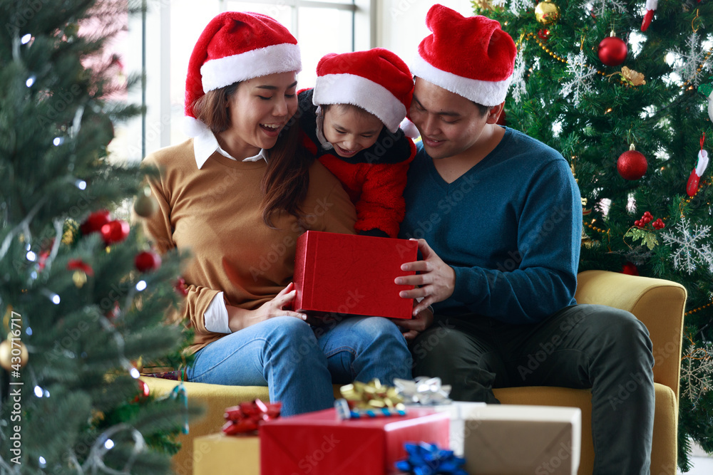 Asian happy family and little girl open magic gift box with Christmas tree in living room that decorated for Christmas festival. People Santa hats sitting On the couch. Concept Christmas gift box.