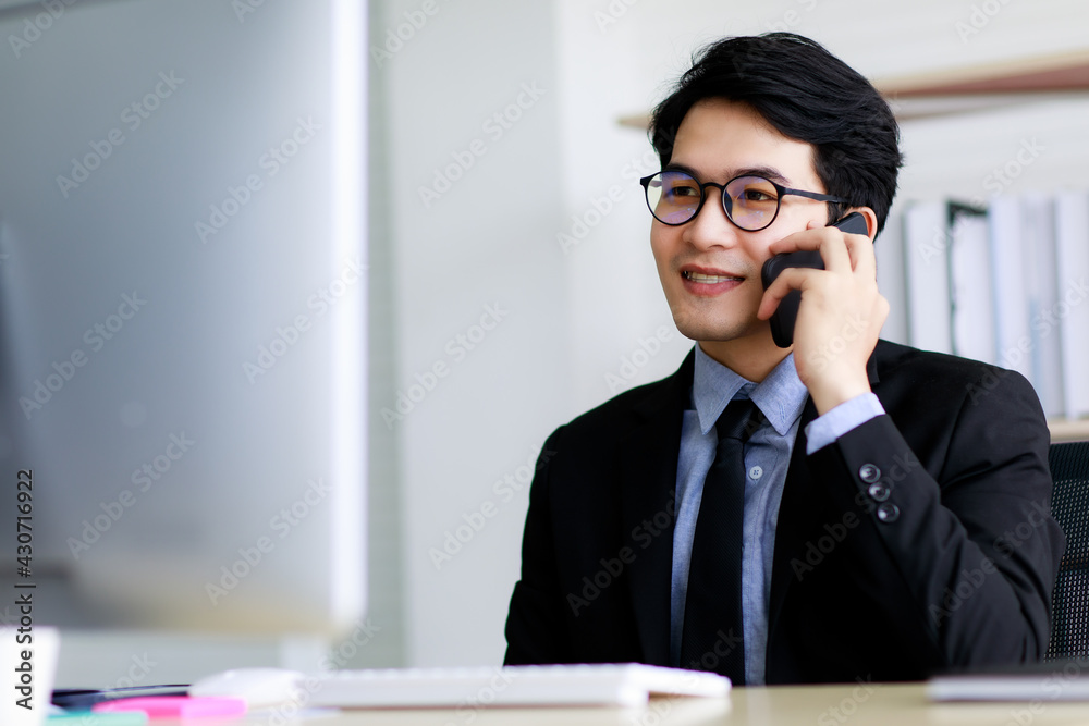 Asian handsome business man wearing eyeglasses, formal black suit with necktie, happily smiling with brace on his teeth while working and talking or communicating on mobile phone in office. Asian handsome business man wearing eyeglasses, formal black suit with necktie, happily smiling with brace on his teeth while working and talking or communicating on mobile phone in office.