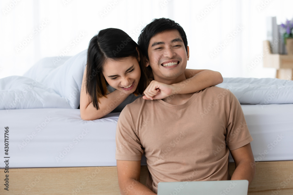 Portrait shot of cute smiling young Asian lover couple in casual clothes relax at home on weekends. The wife lies on the bed behind her husband that sits on the floor and embracing him with happiness