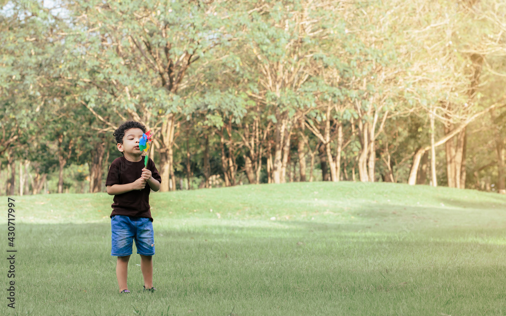 Little african curly hair boy wearing brown shirt playing and holding and blowing wind turbine toy in green field outdoor park. Education and Learning Concept. Little african curly hair boy wearing brown shirt playing and holding and blowing wind turbine toy in green field outdoor park. Education and Learning Concept.