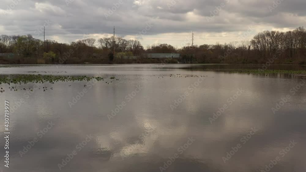 A drone view over a quiet lake, surrounded by dry trees on a cloudy day. The cloudy skies reflect on the smooth water as the camera dolly in over the still water. A drone view over a quiet lake, surrounded by dry trees on a cloudy day. The cloudy skies reflect on the smooth water as the camera dolly in over the still water.