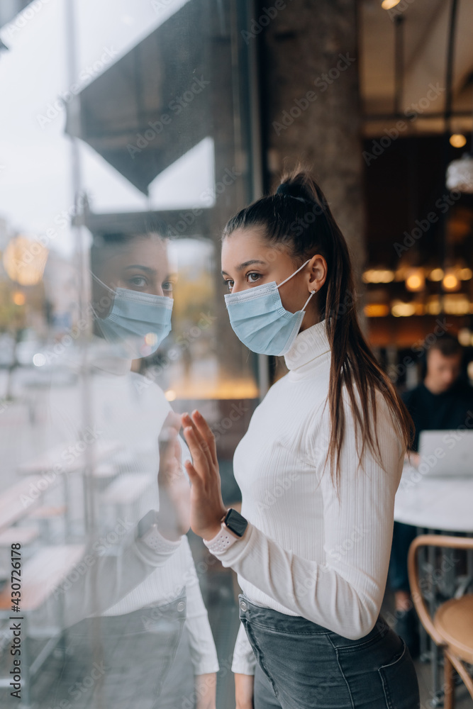Young woman in face mask standing in front of windows in cafe.
