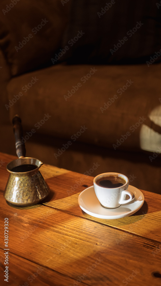Small demitasse cup of Greek coffee on a saucer and a briki sitting on a wooden coffee table with window light illumination