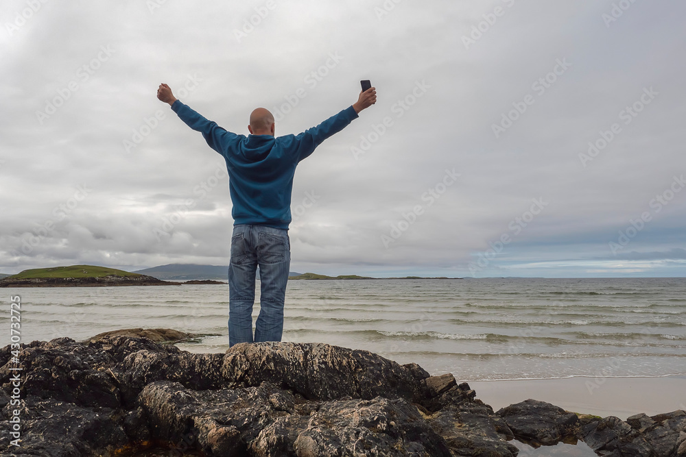 Excited bald man standing on a rock by the ocean, hands up in the air, beautiful and calm scenery in the background. West coast of Ireland. Travel and tourism concept