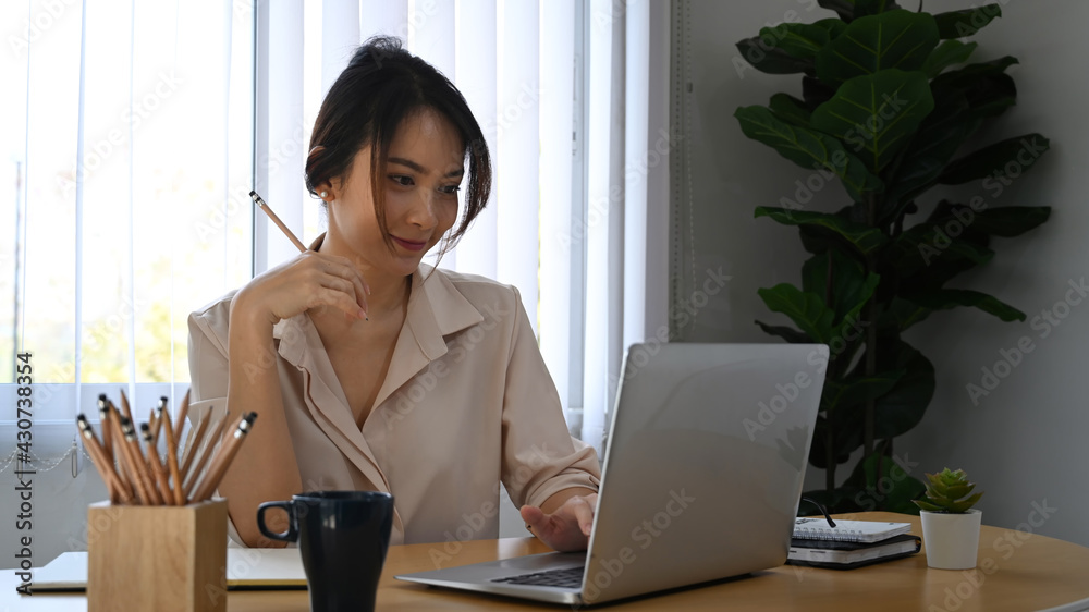 Smiling female employee holding pen and looking at screen of laptop computer. Smiling female employee holding pen and looking at screen of laptop computer.