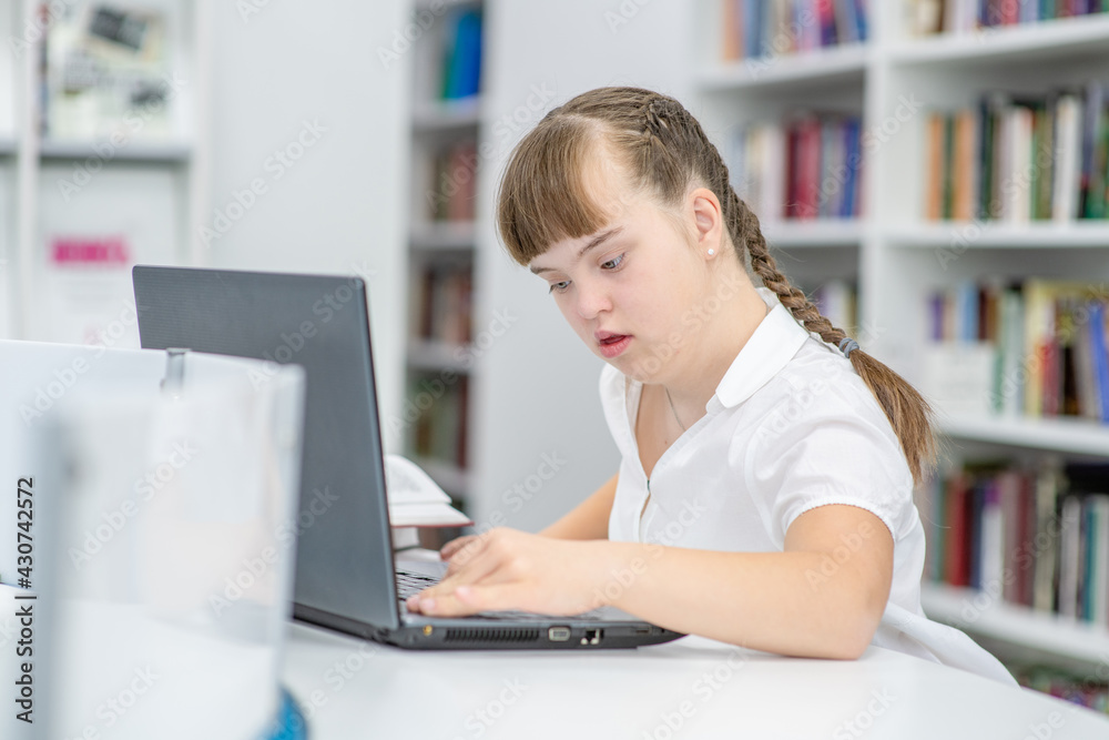 Clever girl with syndrome down uses a laptop at library. Education for disabled children concept Clever girl with syndrome down uses a laptop at library. Education for disabled children concept