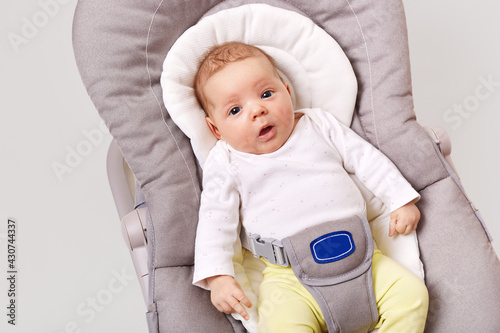 A little newborn girl lies in baby rocking chair, looks around carefully and with interest, exploring the world around her, dressed in a bodysuit and pants, isolated over white background. photo