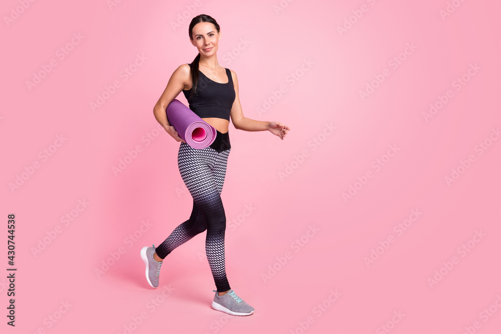 Photo of positive cute young woman dressed sportive outfit walking smiling holding rolled mat empty space isolated pink color background