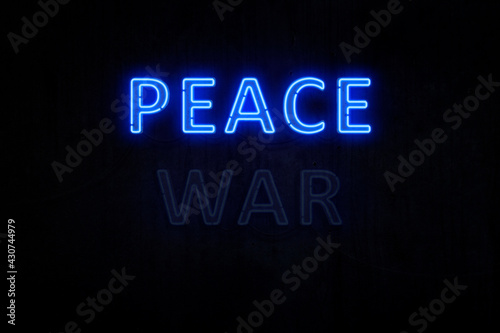 Illuminated and glowing blue "PEACE" neon sign on dark, almost black, grungy wall background. "WAR" neon sign is off. Concept photo of peace and how peace wins over war.