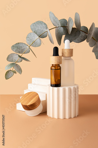 Cosmetic skin care box and two bottles with dropper and wooden covers,from glass staying on the geometrical podiums.Empty space for standing products.Eucalyptus branch above.Pastel beige color.