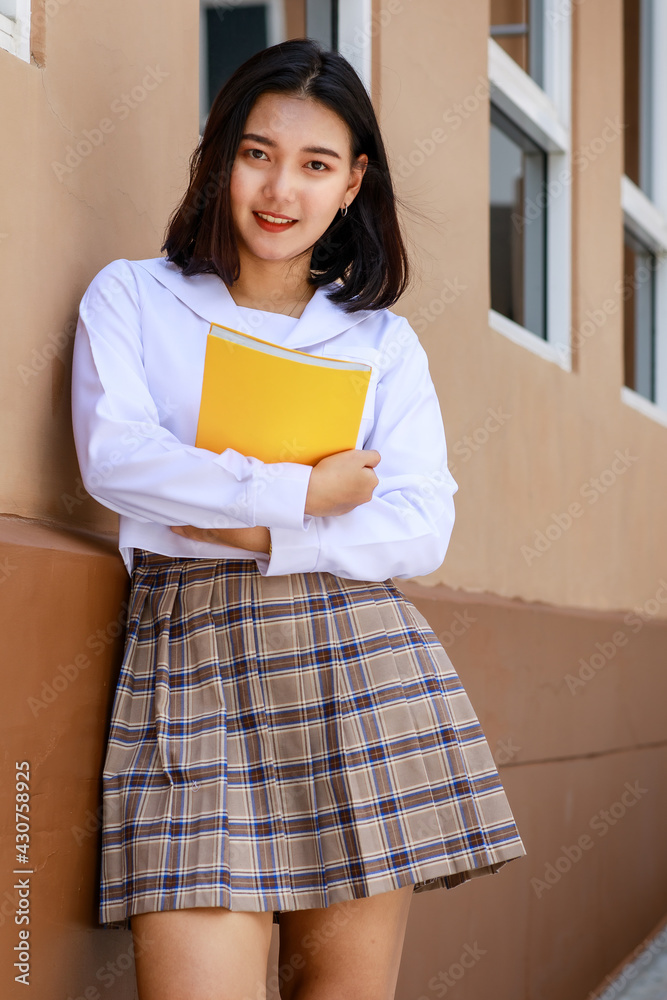 Cute and young girl wearing Japanese, Korean style schoolgirl uniform holding book and pose to camera with fun and happy in front of school building Cute and young girl wearing Japanese, Korean style schoolgirl uniform holding book and pose to camera with fun and happy in front of school building