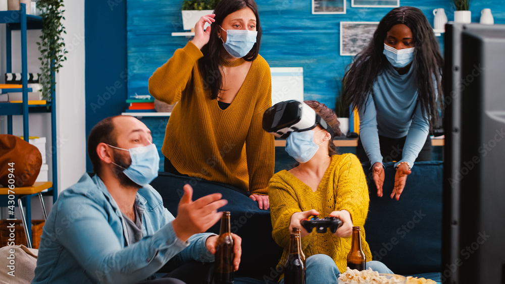 Young woman experiencing virtual reality playing video games with vr headset and joystick in home living room while friends supporting her keeping social distancing because of global pandemic.