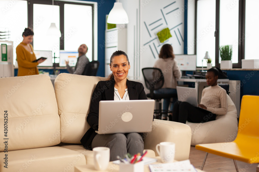 Hispanic business woman smiling at camera sitting on couch typing on laptop while diverse colleagues working in background. Multiethnic coworkers analysing startup financial reports in modern office