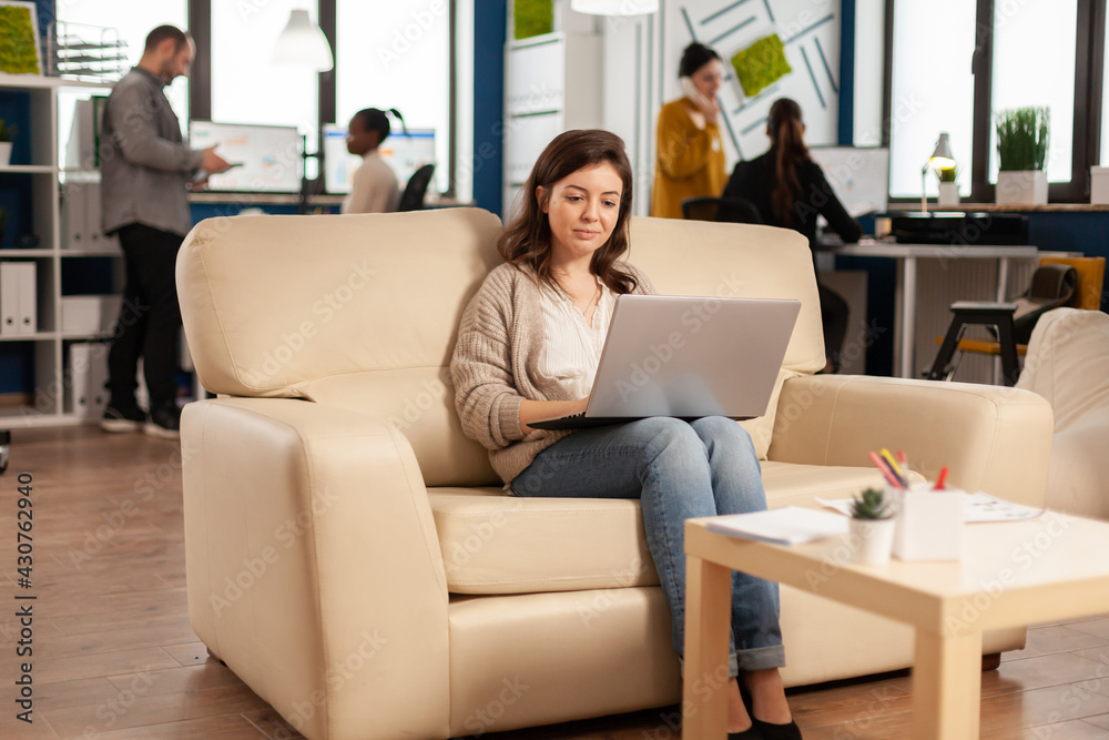 Business woman typing on laptop sitting on couch in start up office while diverse team working in background analysing statistics data. Multiethnic coworkers talking about startup financial company Business woman typing on laptop sitting on couch in start up office while diverse team working in background analysing statistics data. Multiethnic coworkers talking about startup financial company
