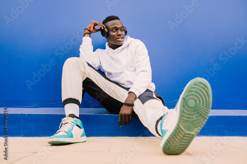 Young afro American black man wearing a white sweatshirt, basketball sneakers and headphones seated on a blue wall looking to the side with confidence photo