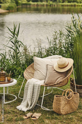 Stylish composition of outdoor garden on the lake with design rattan armchair, coffee table, plaid, pillows, drinks and elegant accessories. Summer chillout mood.