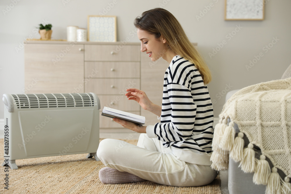 Young woman with book sitting on floor near electric heater at home