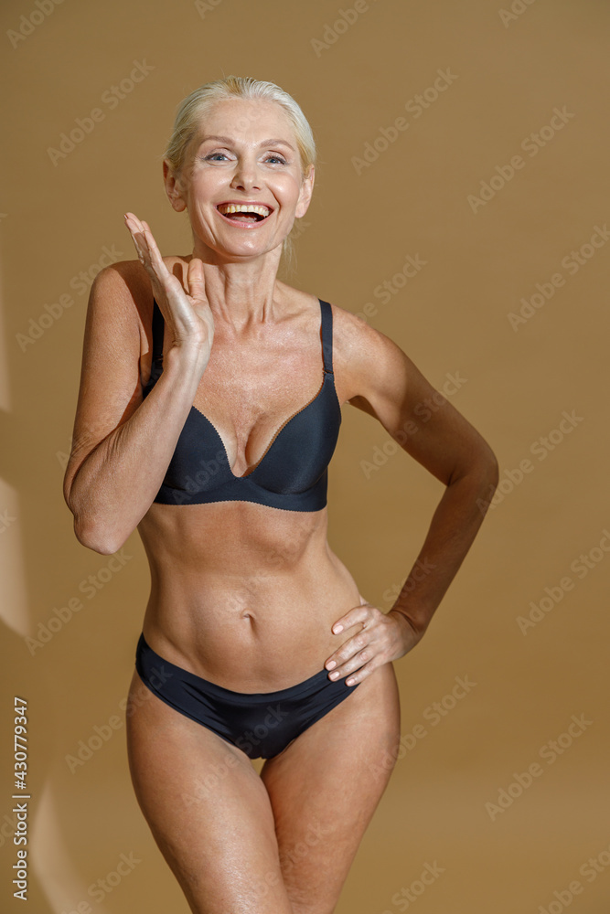 Studio shot of excited mature woman in black underwear smiling at camera  while standing and posing half naked isolated over beige background Photos  | Adobe Stock