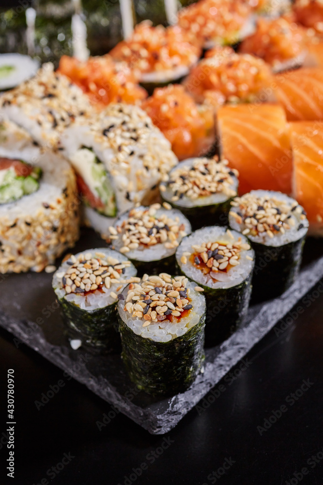 Close up of maki sushi sprinkled with sesame seeds on a stone plate on a dark background. Close up of maki sushi sprinkled with sesame seeds on a stone plate on a dark background.