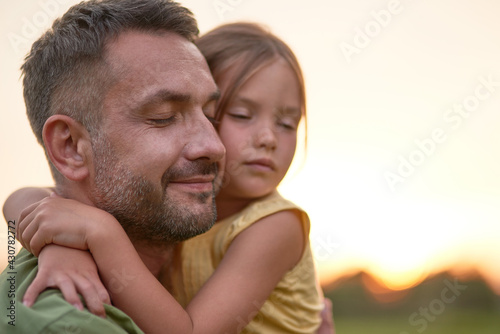 Close up shot of young father and adorable little daughter embracing each other, posing with eyes closed while spending time outdoors in the park on a summer day © Kostiantyn