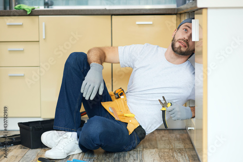 Get rid of leaks. Full length shot of young handyman, plumber wearing tool belt examining a sink pipe before fixing it in the kitchen