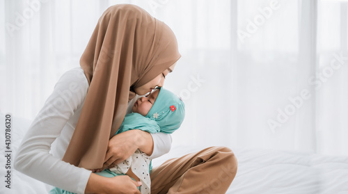 Happy asian muslim mother holding and kissing adorable little baby daughter wearing hijab in her arms on white bed in bedroom.