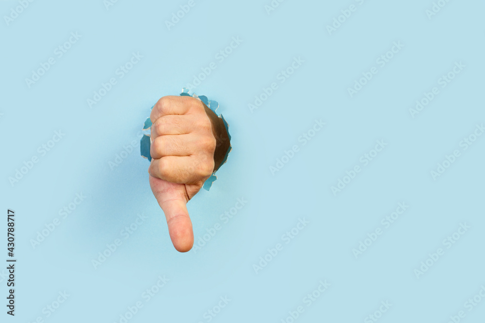 Mano de hombre a través de un agujero en un papel celeste roto haciendo el gesto con el dedo pulgar hacia abajo. Vista de frente. Copy space Mano de hombre a través de un agujero en un papel celeste roto haciendo el gesto con el dedo pulgar hacia abajo. Vista de frente. Copy space