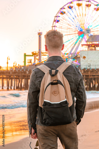 View from the back of a young blonde man with a backpack on Santa Monica beach against the background of an orange sunset in Los Angeles, California