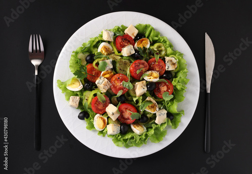 Healthy salad of cherry tomatoes, cucumbers, peppers, black olives, quail eggs and gorgonzolla cheese on black background, top view