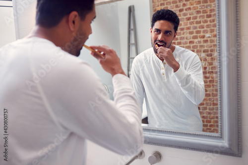 Man Wearing Pyjamas At Home In Modern Bathroom Brushing Teeth In Mirror