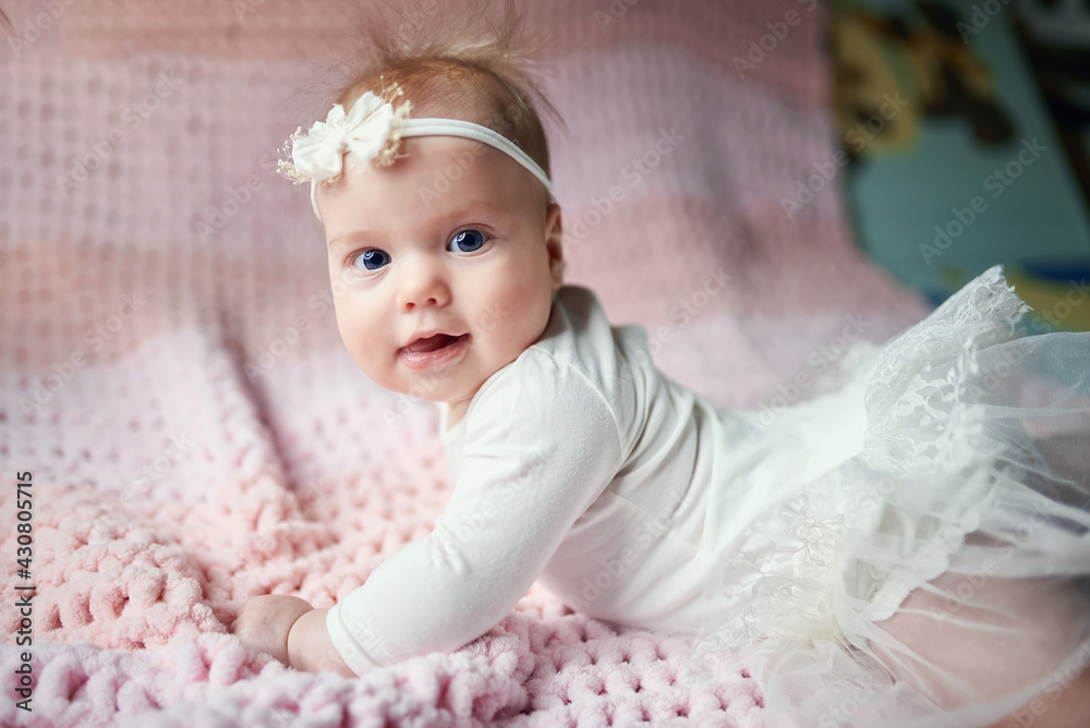 a cute baby in a fluffy dress looks into the camera with a banter on his head and in white clothes. The mouth is open and the tongue is visible. Blue eyes and fluffy hair