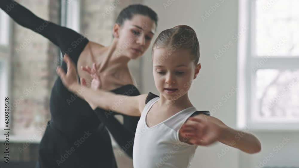 Tracking waist-up shot of young professional ballerina in black ballet dress dancing together with pretty little girl, moving synchronically at dance class