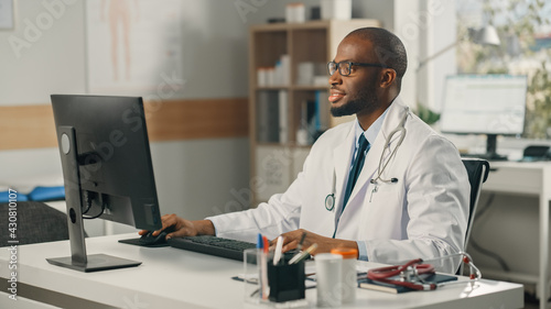 Experienced African American Male Doctor Wearing White Coat Working on Personal Computer at His Office. Medical Health Care Professional Working with Test Results, Patient Treatment Planning.