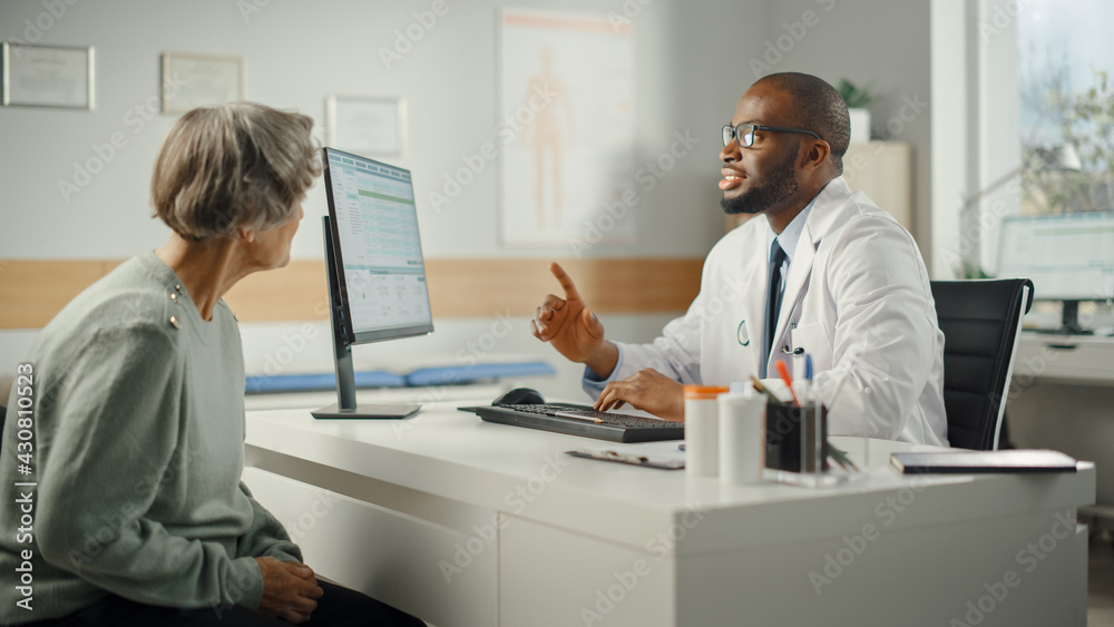 African American Family Doctor is Prescribing Medication to Senior Female Patient and Speaking with Her During Consultation in a Health Clinic. Physician Sitting Behind a Desk in Hospital Office.