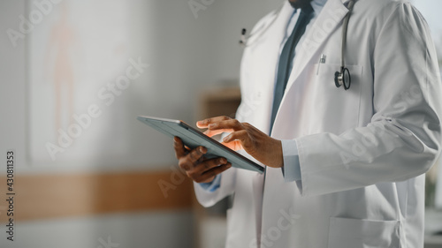 Close Up Shot of a African American Male Doctor Wearing White Coat Working on Tablet Computer at His Office. Medical Health Care Professional Working with Test Results, Patient Treatment Planning.