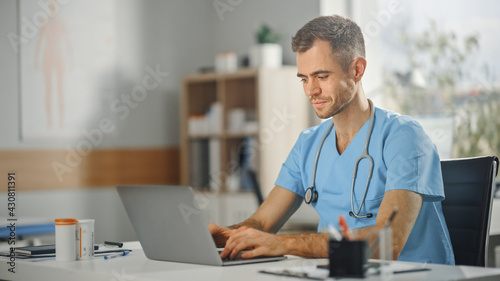 Experienced Male Nurse Wearing Blue Uniform Working on Laptop Computer at Doctor's Office. Medical Health Care Professional Working On Battling Stereotypes to Gender Diversity in Nursing Career. photo