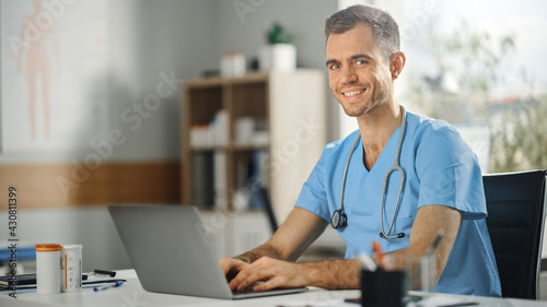 Male Nurse Wearing Blue Uniform Working on Laptop Computer at Doctor's Office and Smiling on Camera. Health Care Professional Working On Battling Stereotypes to Gender Diversity in Nursing Career. photo