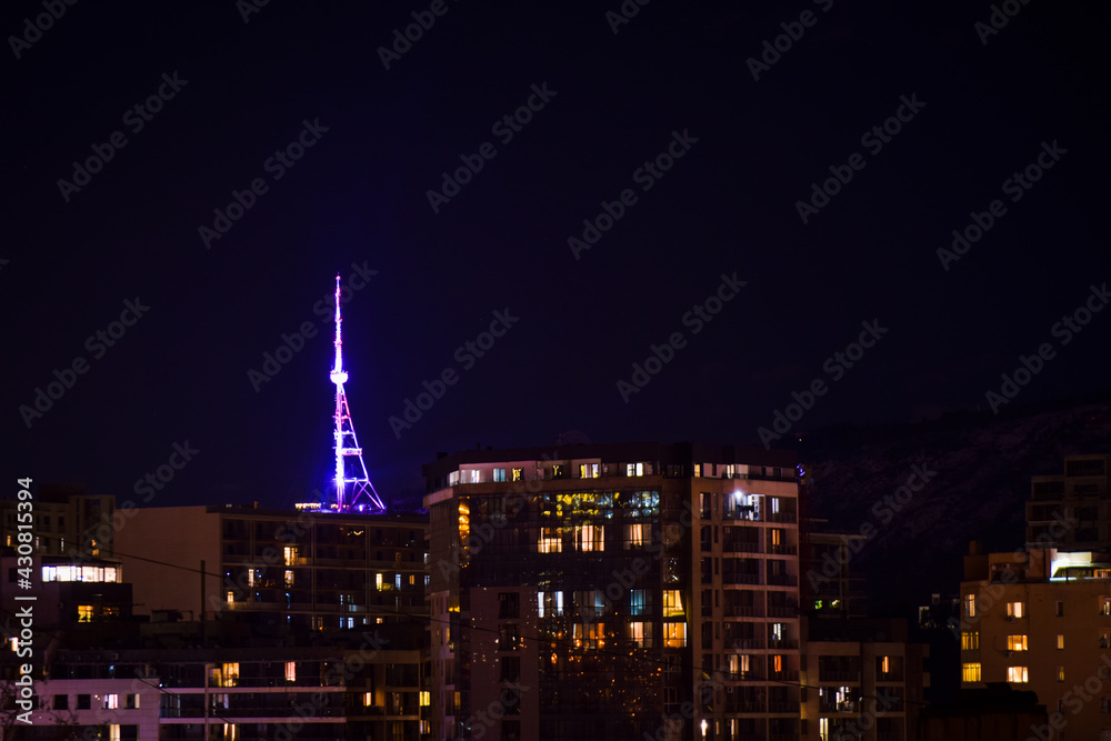 Naklejka premium Tbilisi, Georgia - 17th april, 2021: Tbilisi TV communication tower illuminated at night. View Georgia capital and house buildings at night concept background.