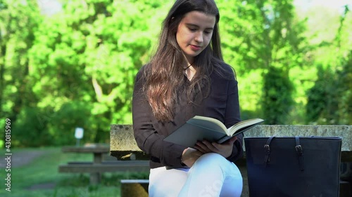 long-haired girl, black shirt and white pants reading a book in nature, sitting at a stone table with a coffee and her bag. reading with background trees and green book teenager studying in the park