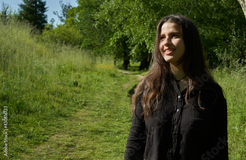 beautiful long-haired woman in black shirt, white pants and brown bag walking through nature with trees in the background and blue sky. teenager walking in the park watching the camera