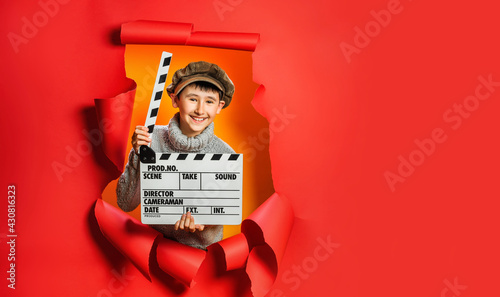 Fotografia Little boy in knitted sweater and retro cap is playing movie maker holding wooden director's clapper board