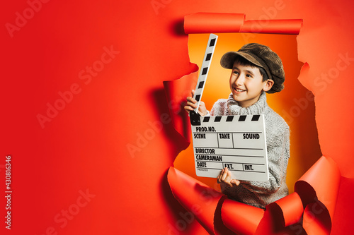 Canvas-taulu Little boy in knitted sweater and retro cap is playing film director holding wooden director's clapper board